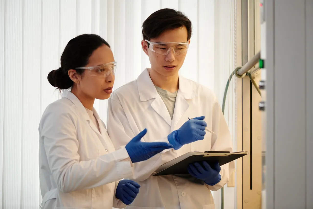 Two lab specialists in safety glasses reviewing results on a clipboard